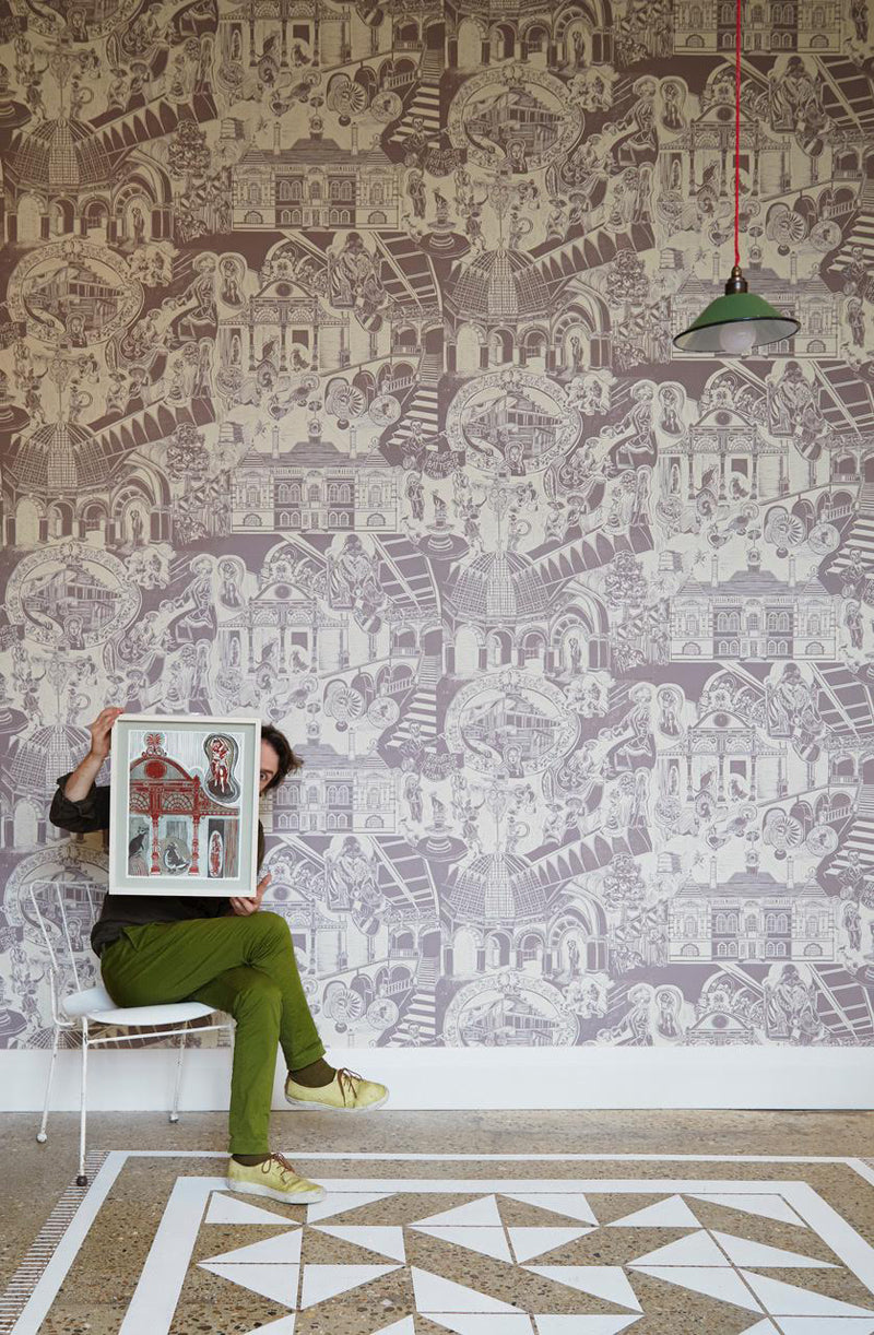 an artist sits on a chair holding one of his prints, in front of a wallpapered wall