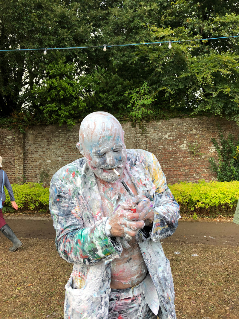 Man lighting his cigarette at Port Eliot festival wearing a suit covered in white and multicoloured paint 